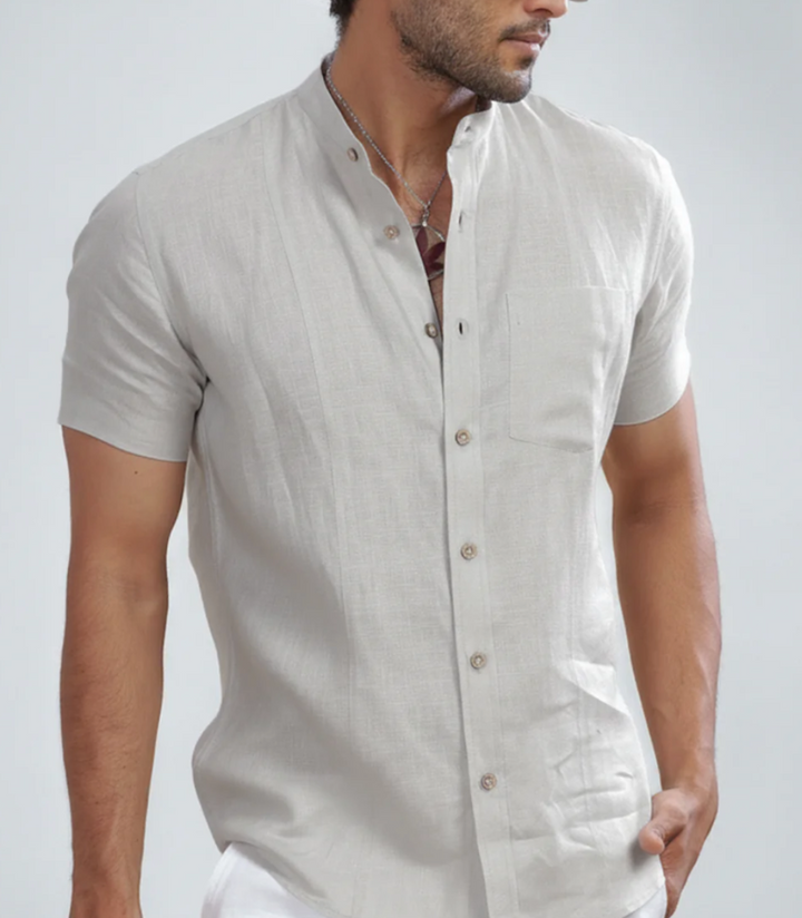 A man wearing a grey  beige short-sleeve linen shirt with a mandarin collar and button-down front, styled with white trousers and dark sunglasses against a light white background.