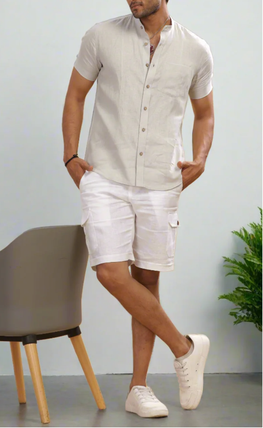 A man modeling a light grey short-sleeve linen shirt and white cargo shorts, standing next to a gray chair in a minimalist room.